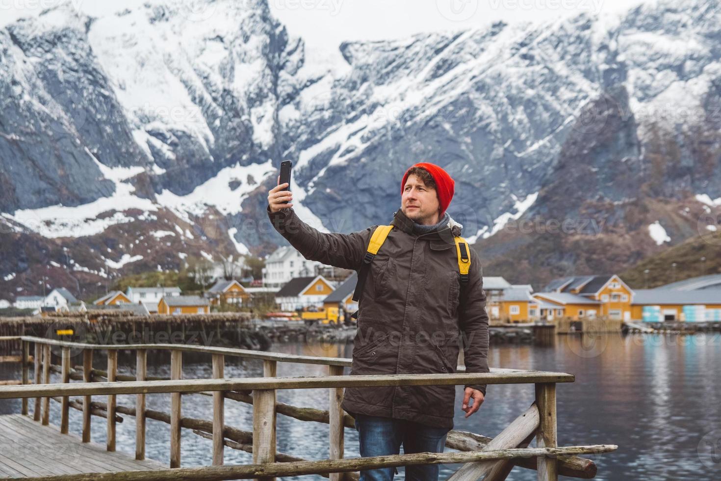 Traveler man taking self-portrait with a smartphone against the background of snowy mountains, rocks and lakes standing on a wooden pier. Place for text or advertising photo