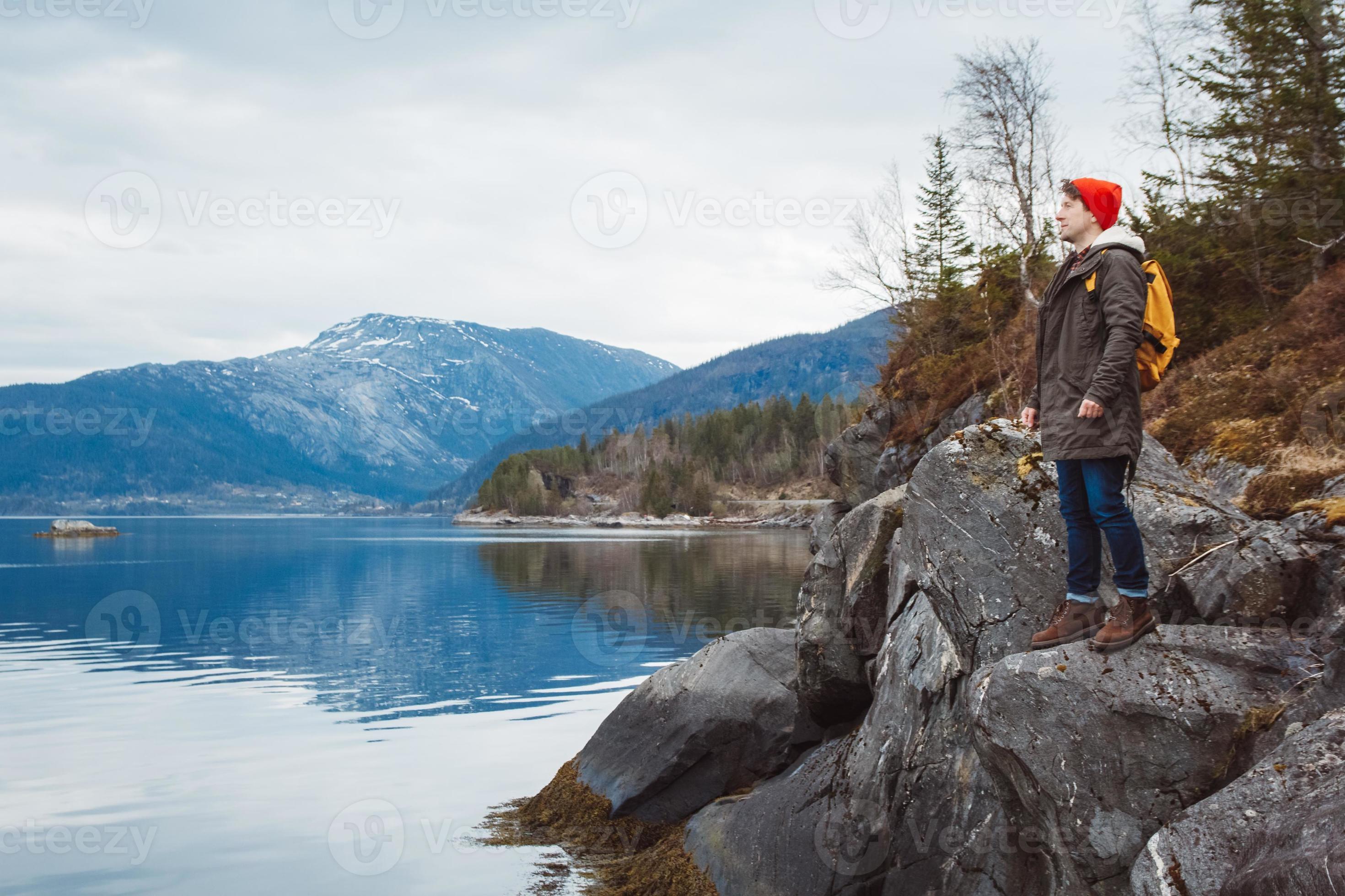Young man with a yellow backpack wearing a red hat standing on a rock on the background of mountain and lake. Space for your text message or promotional content. Travel lifestyle concept photo