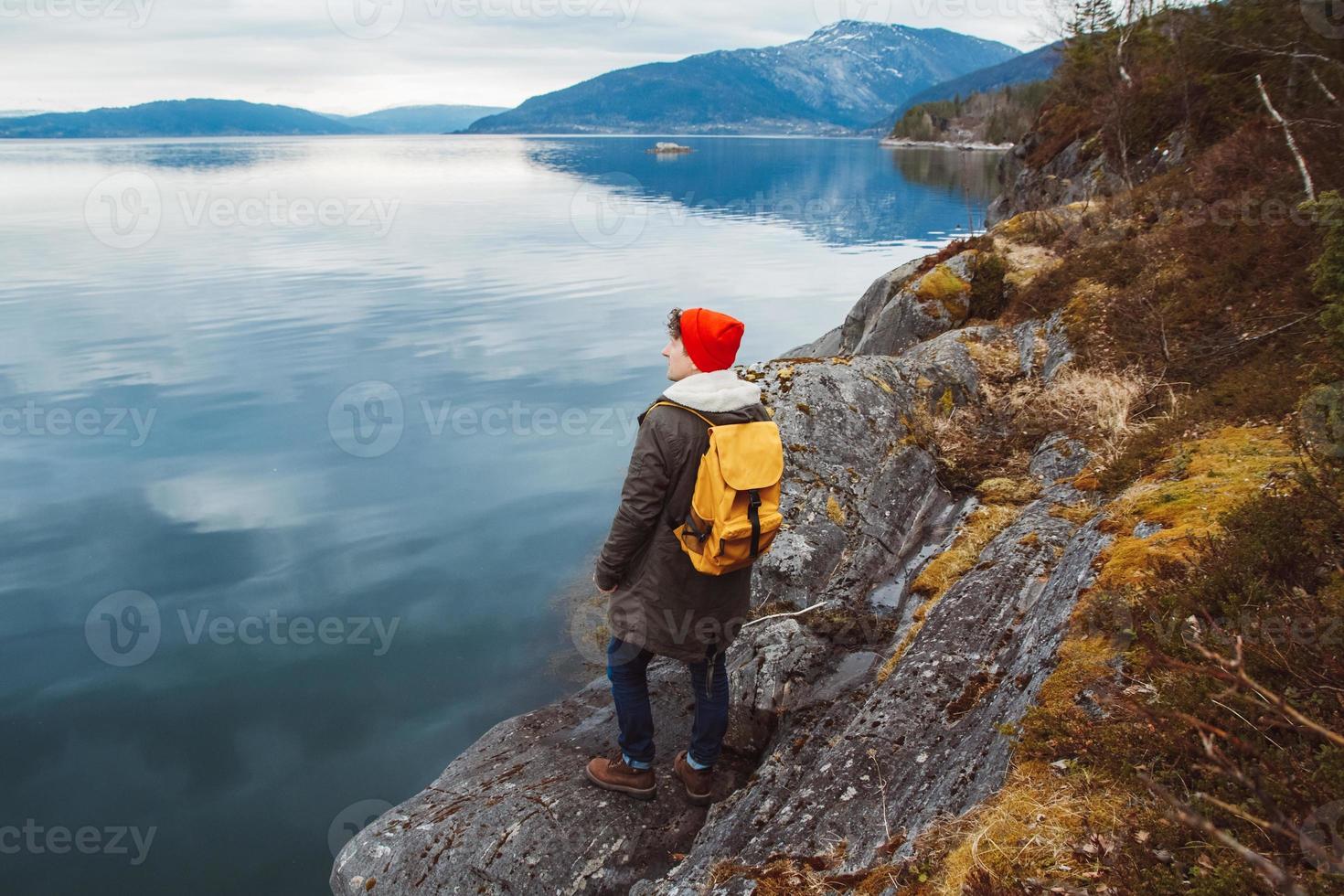 joven con una mochila amarilla con un sombrero rojo de pie sobre una roca en el fondo de la montaña y el lago. espacio para su mensaje de texto o contenido promocional. concepto de estilo de vida de viaje foto