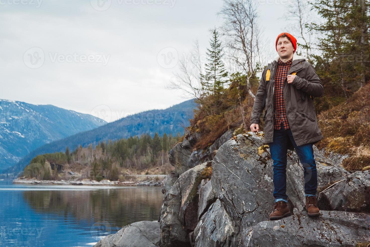 joven con una mochila amarilla con un sombrero rojo de pie sobre una roca en el fondo de la montaña y el lago. espacio para su mensaje de texto o contenido promocional. concepto de estilo de vida de viaje foto