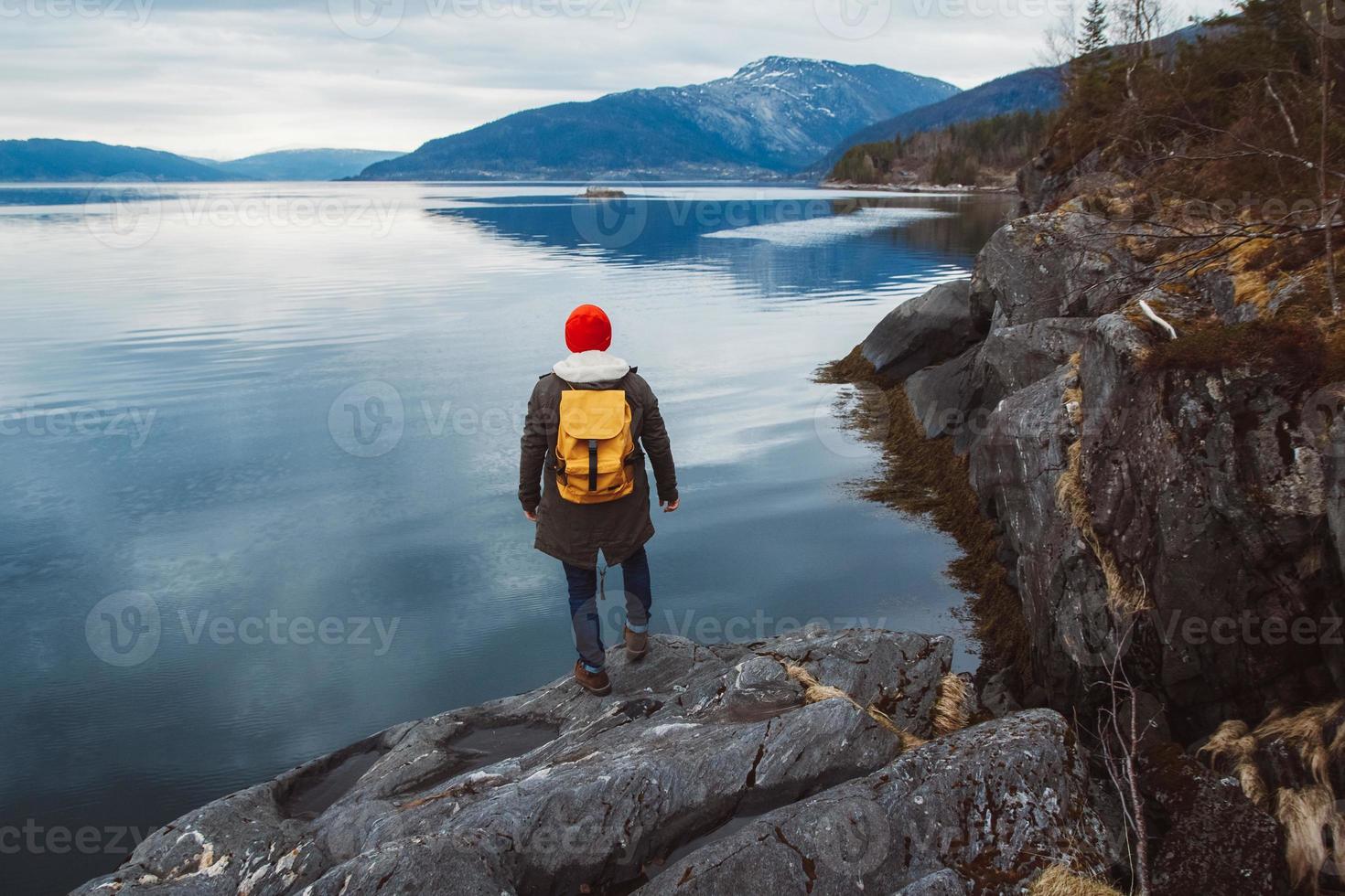 joven con una mochila amarilla con un sombrero rojo de pie sobre una roca en el fondo de la montaña y el lago. espacio para su mensaje de texto o contenido promocional. concepto de estilo de vida de viaje foto