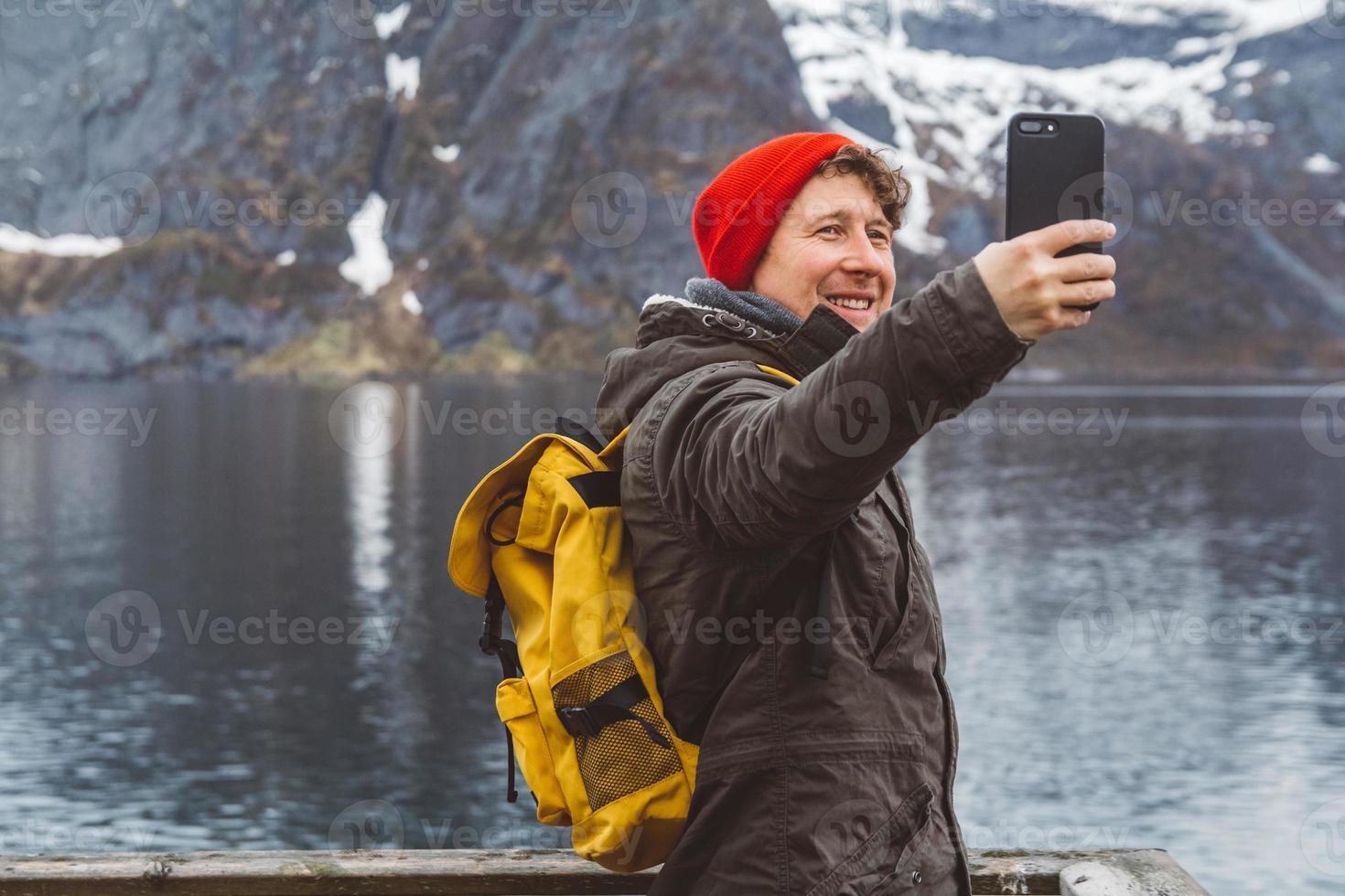 Traveler man taking self-portrait with a smartphone against the background of snowy mountains, rocks and lakes standing on a wooden pier. Place for text or advertising photo