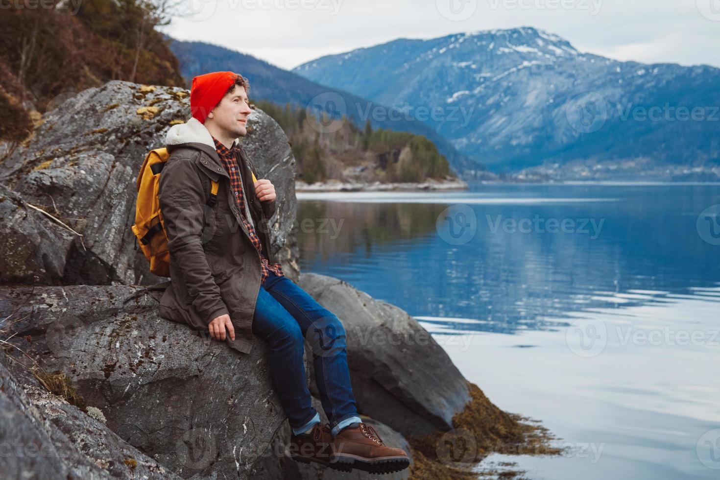 Young man with a yellow backpack wearing a red hat sitting on the shore on the background of mountain and lake. Space for your text message or promotional content. Travel lifestyle concept. photo