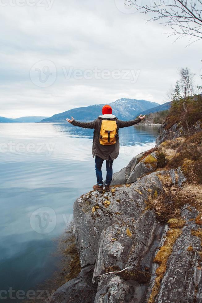 Young man with a yellow backpack wearing a red hat standing on a rock on the background of mountain and lake. Space for your text message or promotional content. Travel lifestyle concept photo