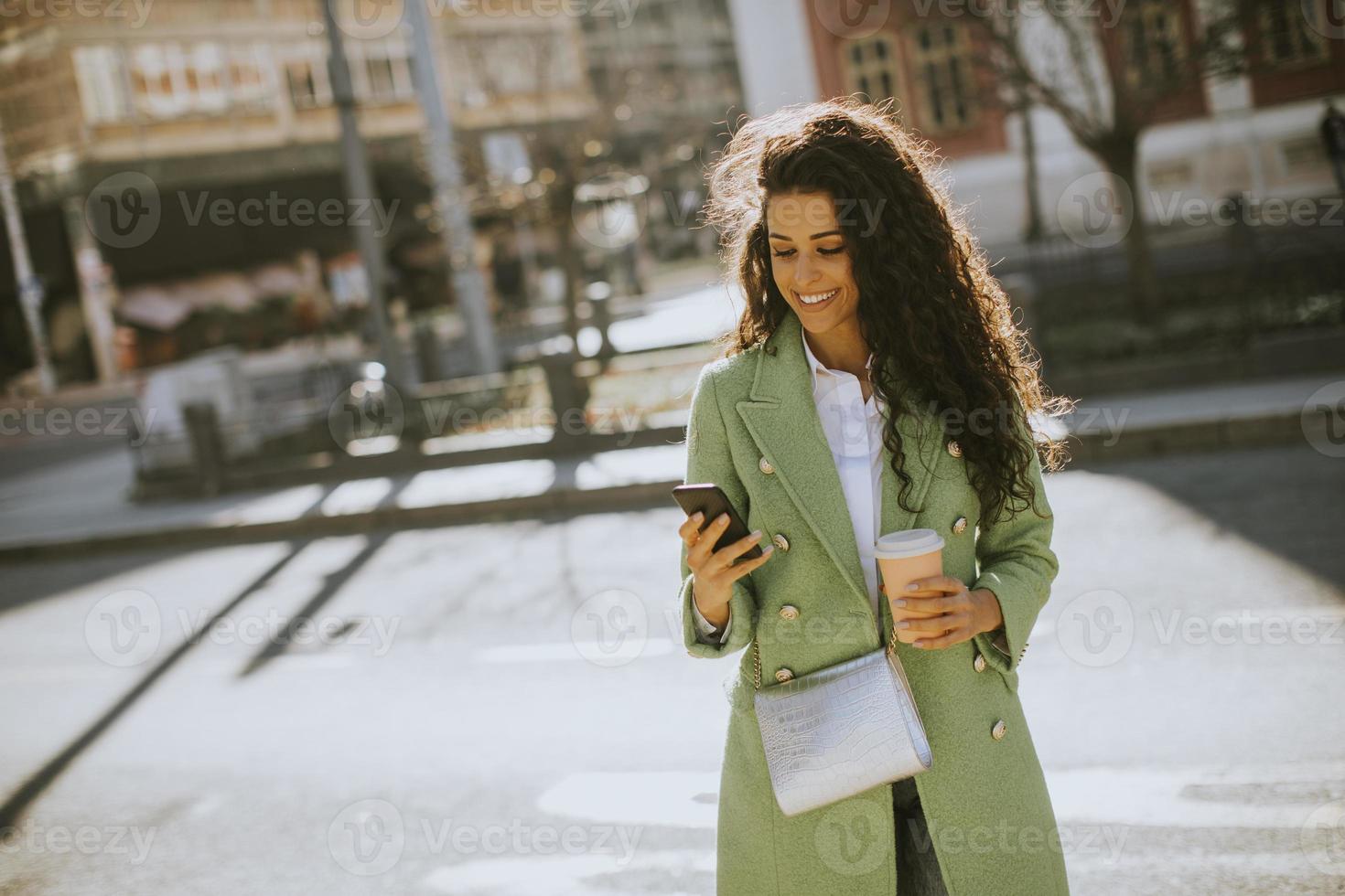 mujer joven usando un teléfono inteligente en la calle y sosteniendo café para llevar foto