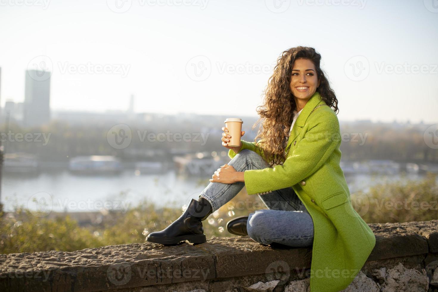 Bastante joven disfrutando del sol de otoño mientras está sentado junto al río y bebiendo café para llevar foto