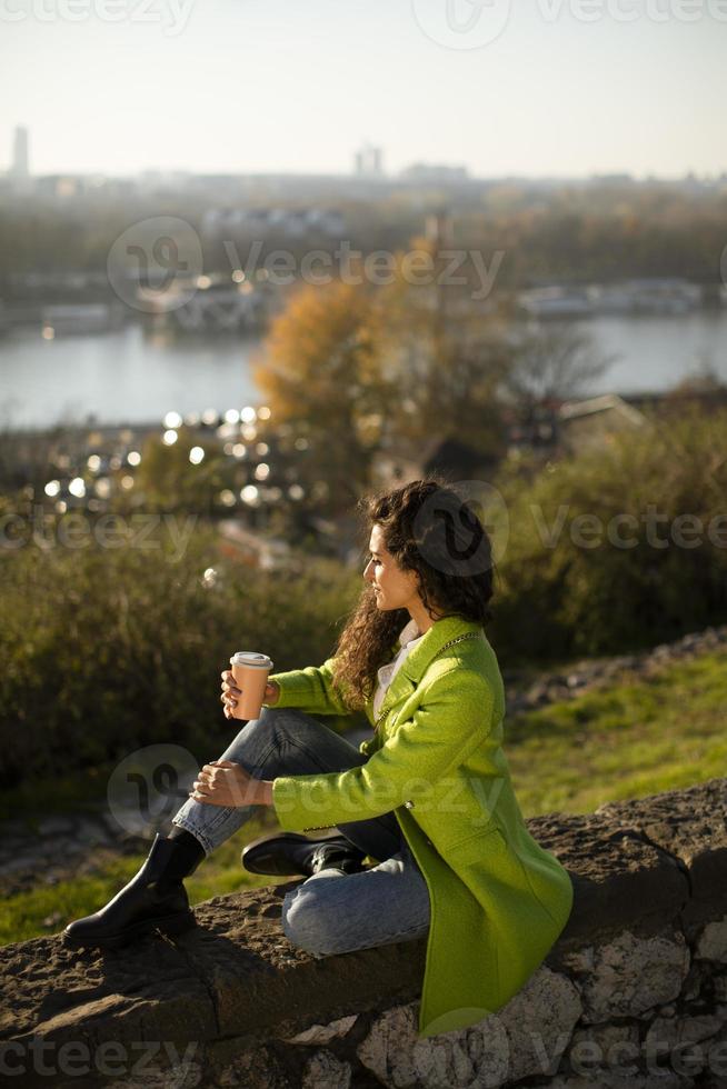 Bastante joven disfrutando del sol de otoño mientras está sentado junto al río y bebiendo café para llevar foto