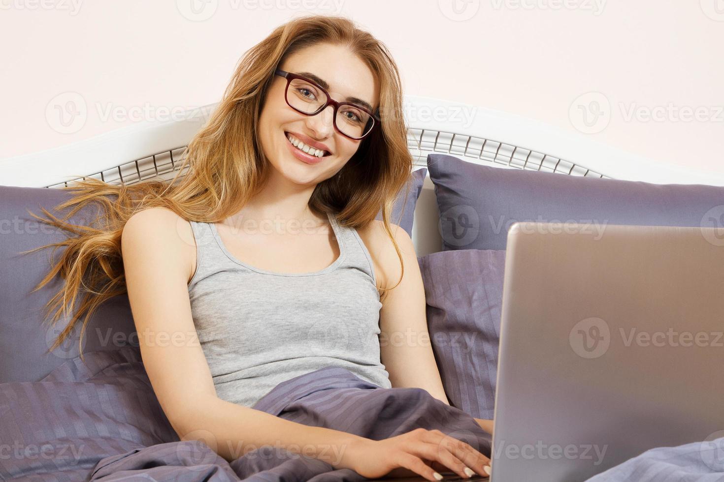 hermosa chica con anteojos acostada en la cama con laptop, feliz casual hermosa mujer trabajando en una laptop sentada en la cama en el dormitorio de la casa. mujer que trabaja en la computadora portátil. ventas digitales foto