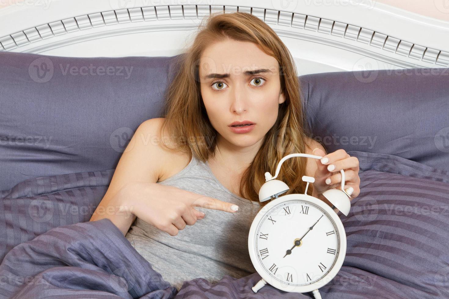 Shocked young woman waking up with alarm, girl point on clocks in bed photo