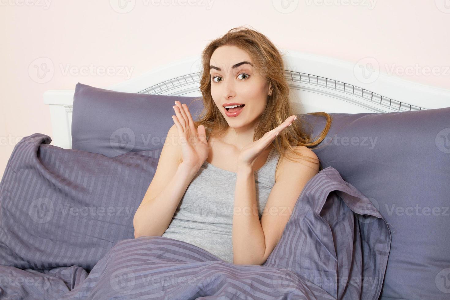 encantadora rubia posando en un dormitorio, mujer positiva con almohada sentada en la cama y sonriendo a la cámara en casa foto