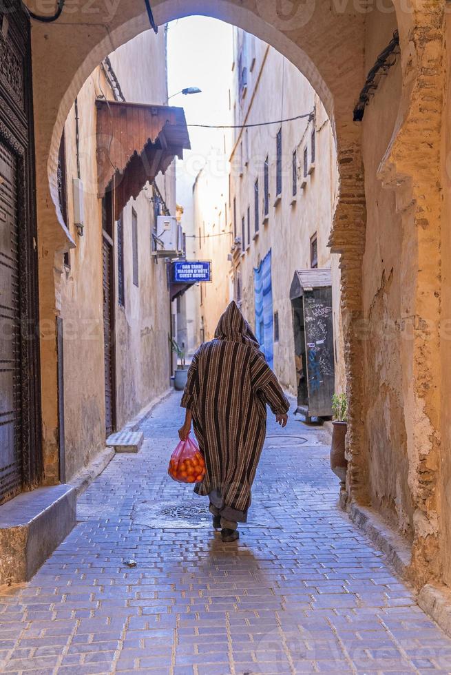 Rear view of man walking in traditional hood clothing while carrying oranges photo