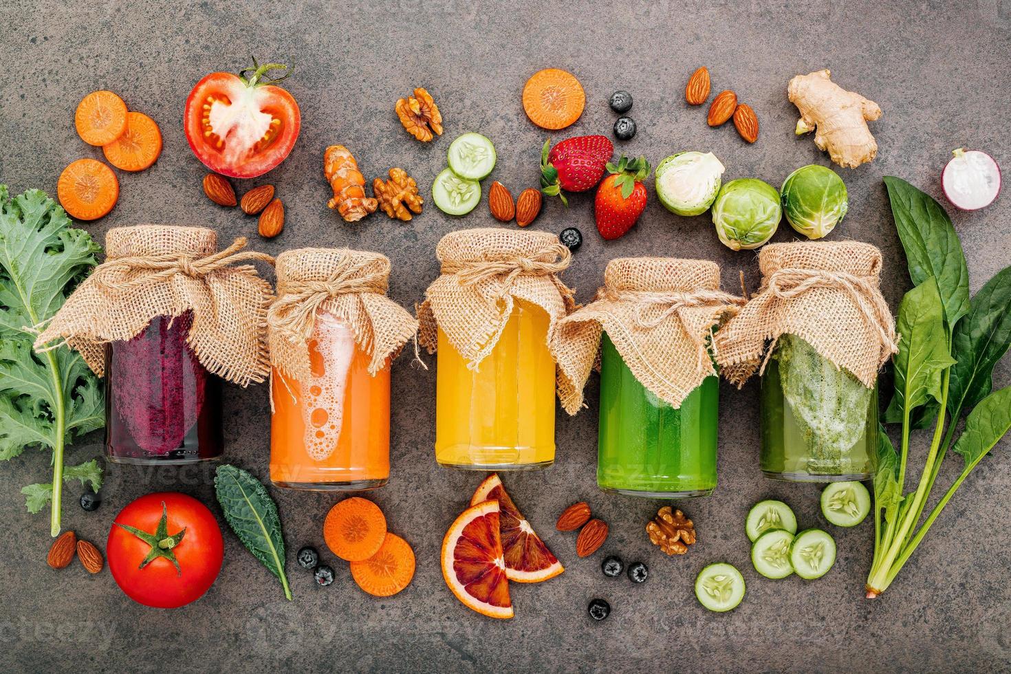 coloridos batidos y jugos saludables en botellas con frutas tropicales frescas y superalimentos sobre fondo de piedra oscura con espacio de copia. foto