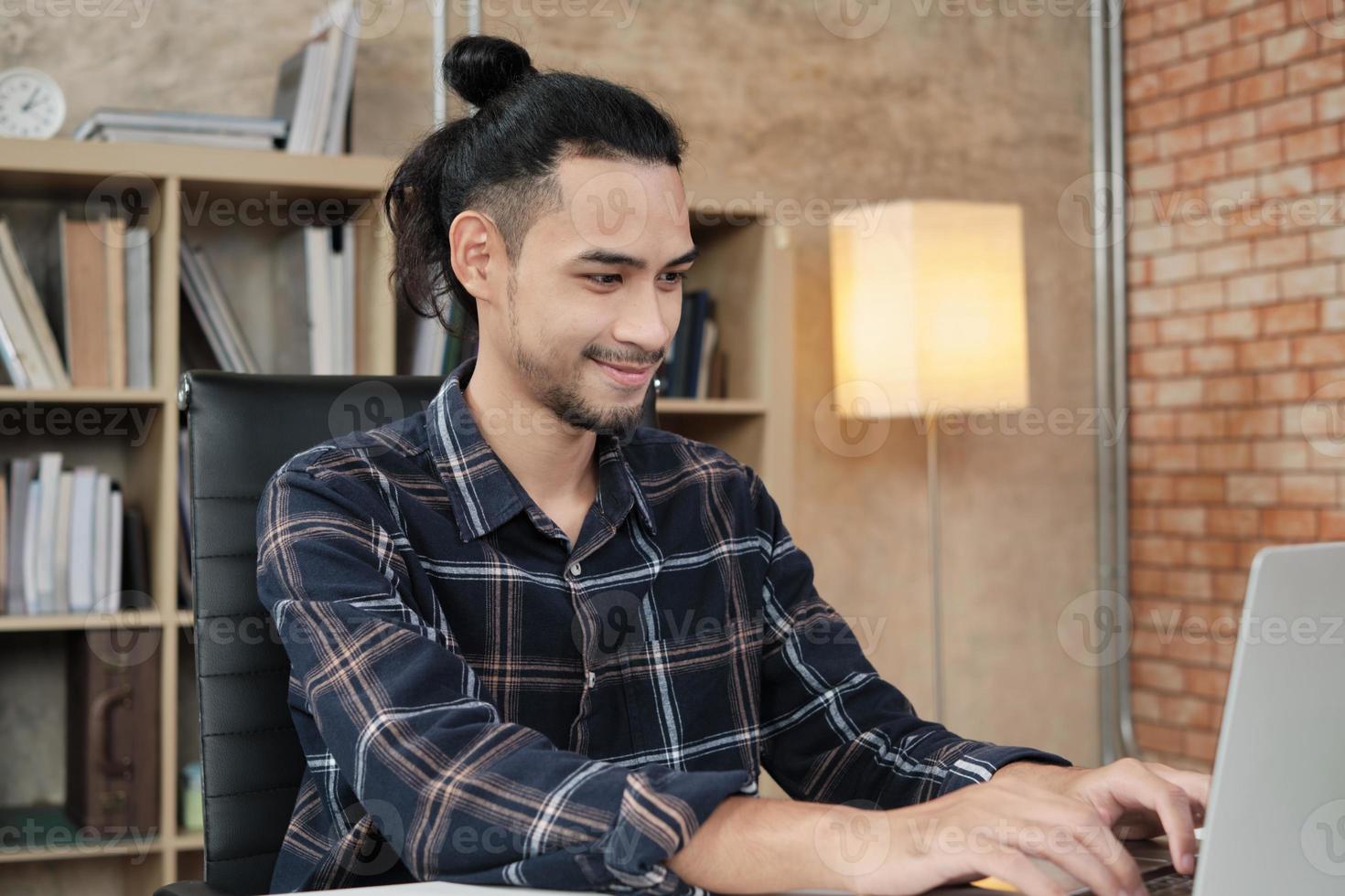 un joven trabajador de etnia asiática usa una computadora portátil para hacer un trabajo creativo en un escritorio blanco frente a la estantería de un lugar de trabajo informal, una persona de negocios nueva y una ocupación de comercio electrónico en línea. foto