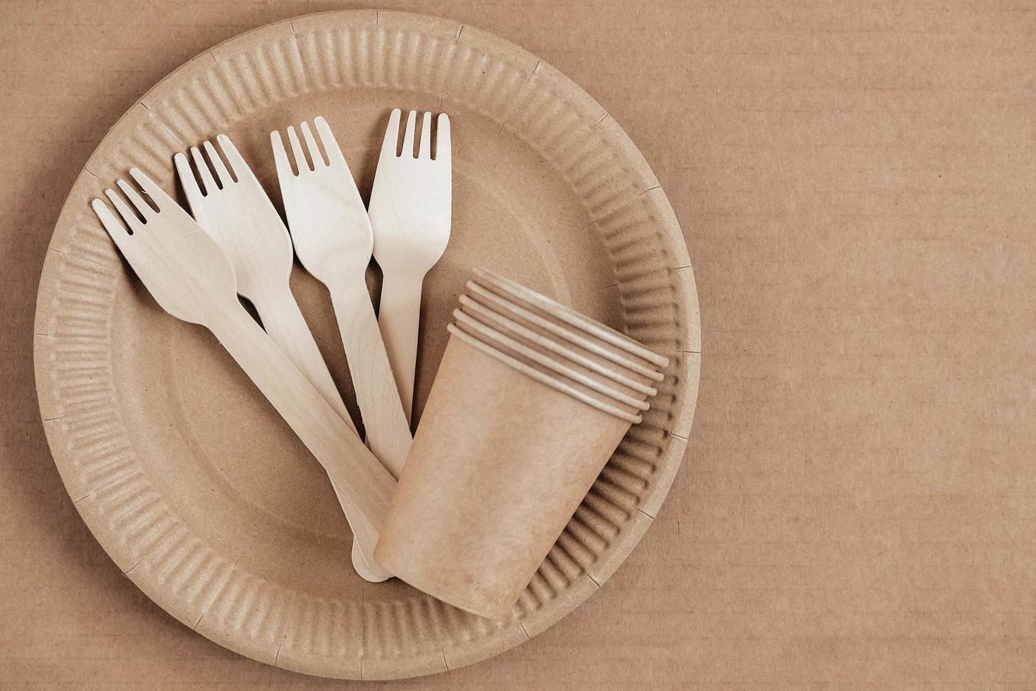 Wooden forks and paper cups with plates on kraft paper background. Eco friendly disposable tableware. Also used in fast food, restaurants, takeaways, picnics. Top view. Copy, empty space for text photo