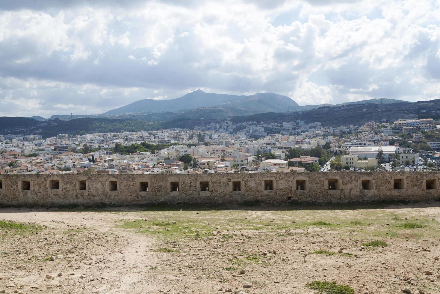 View of resort Greek architecture Rethymno city-port, built by Venetians, from height of Fortezza Castle - fortress on hill Paleokastro. Red tiled roofs and mountains in background. Crete. photo