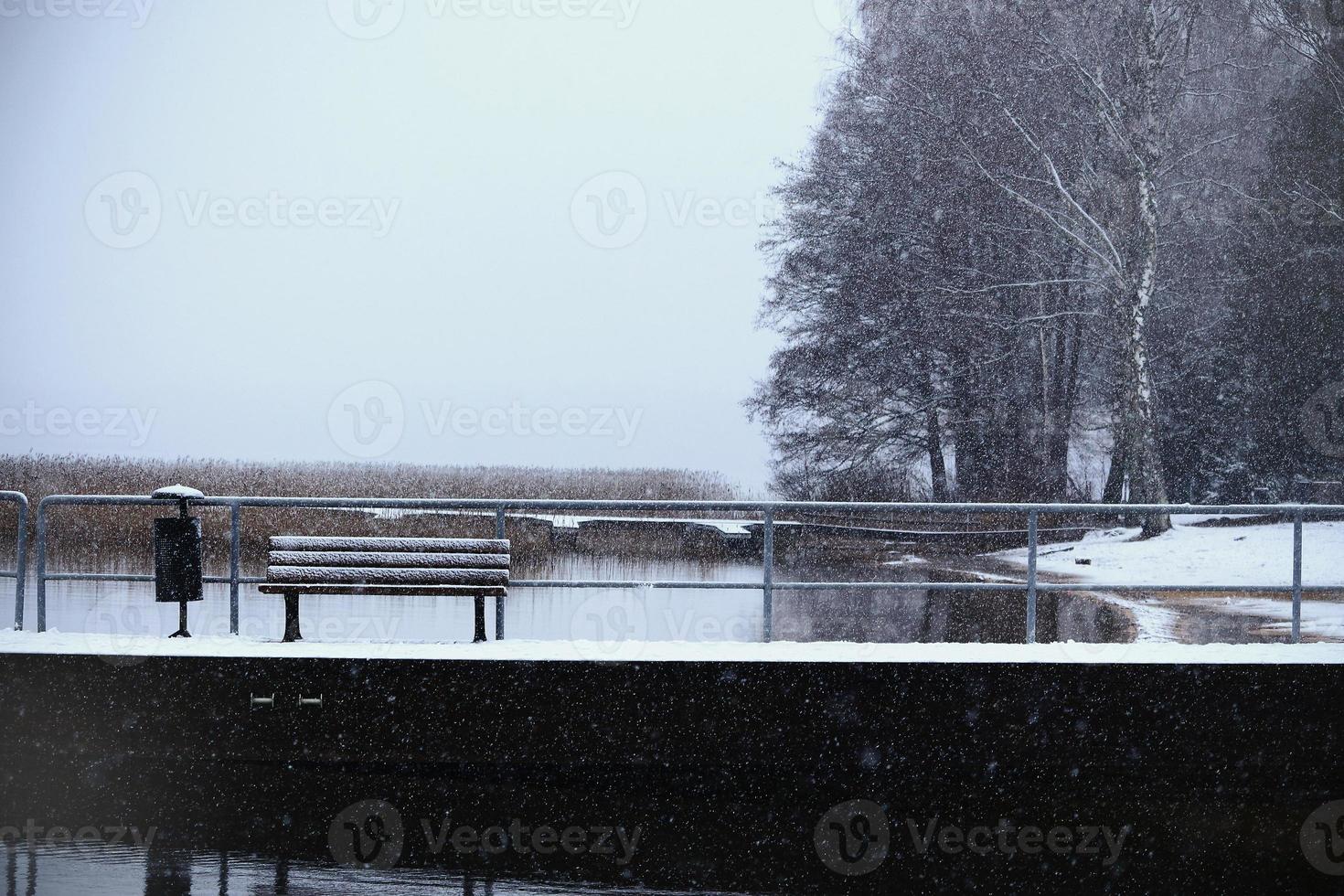 Winter lake landscape with forest along the shore and bench with garbage bin standing on pier photo