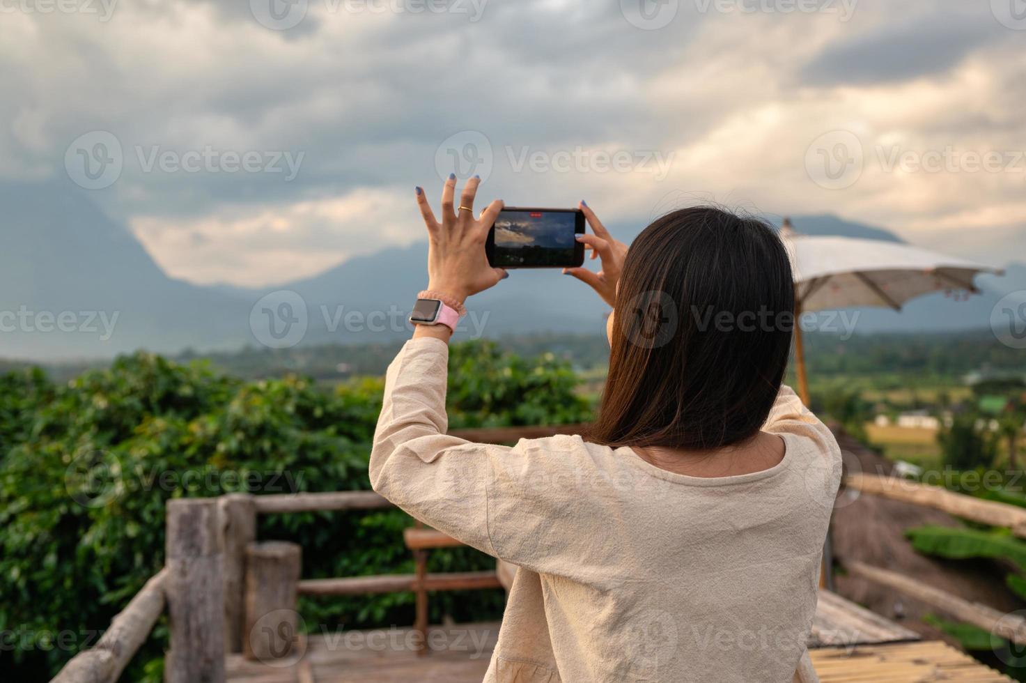 Asian woman taking a video of mountain view by smartphone on wooden balcony in vacation at evening photo