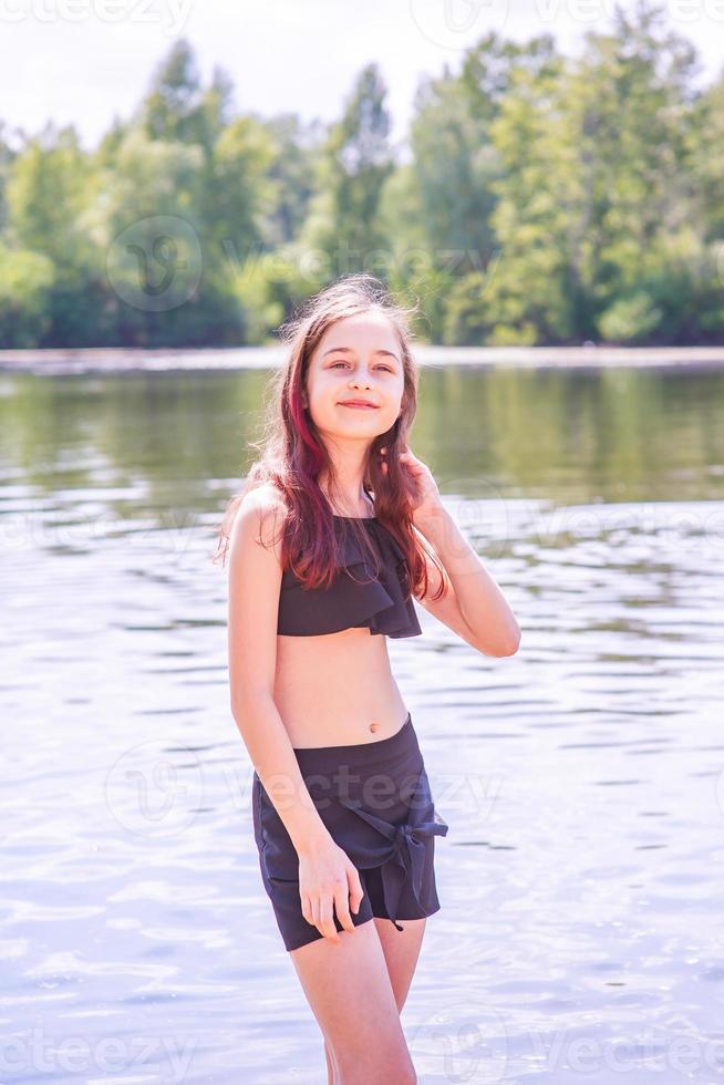 A teenage girl by the river in a black swimsuit and black shorts in summer. Children and rest. photo
