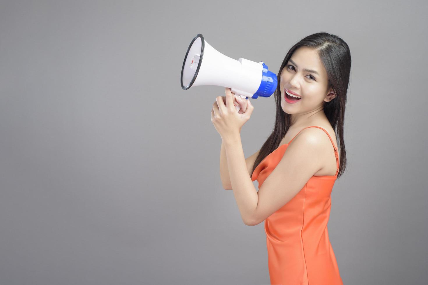 Fashion portrait of beautiful woman wearing orange dress is using megaphone isolated over gray background studio photo