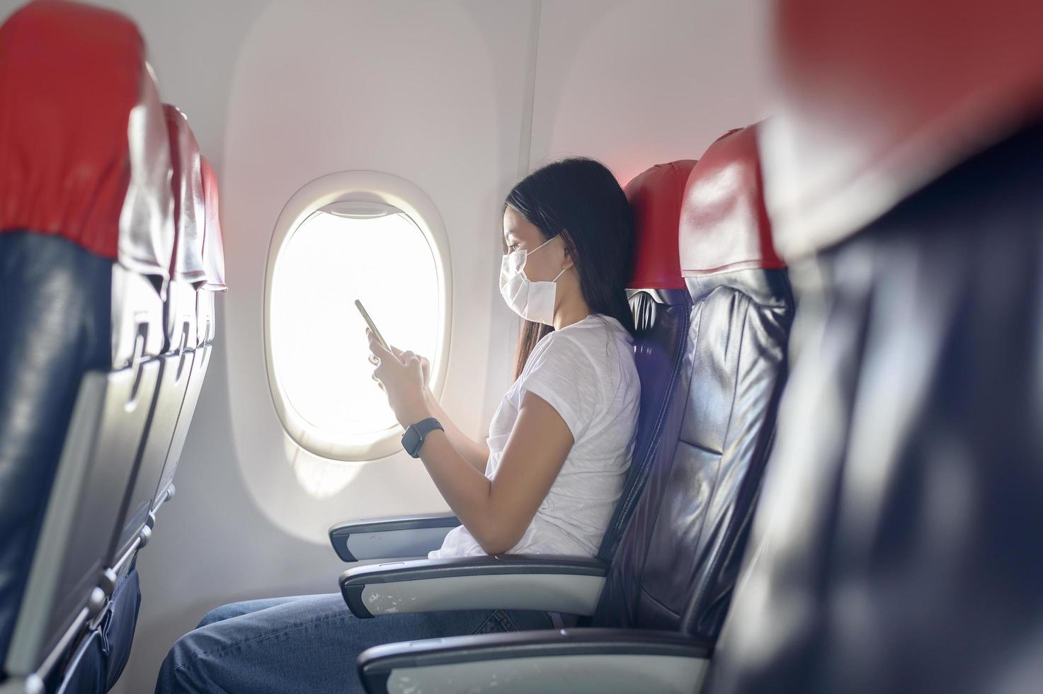 mujer viajera con máscara protectora a bordo del avión usando un teléfono inteligente, viaje bajo la pandemia de covid-19, viajes de seguridad, protocolo de distanciamiento social, nuevo concepto de viaje normal foto