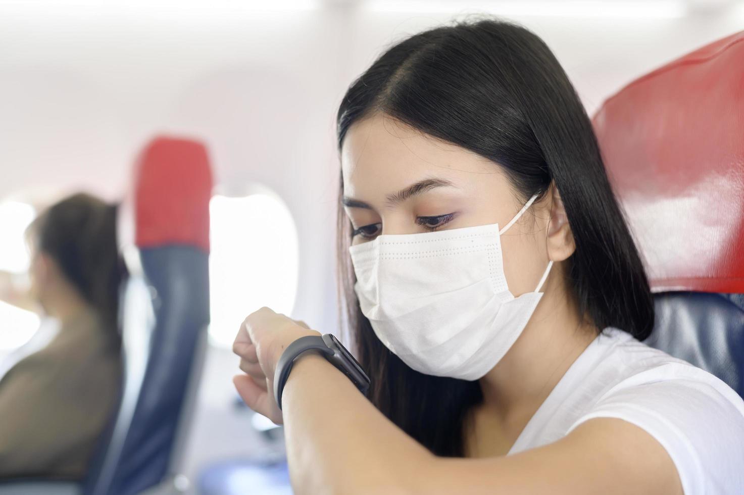 mujer viajera con máscara protectora a bordo del avión usando reloj inteligente, viaje bajo la pandemia de covid-19, viajes de seguridad, protocolo de distanciamiento social, nuevo concepto de viaje normal foto