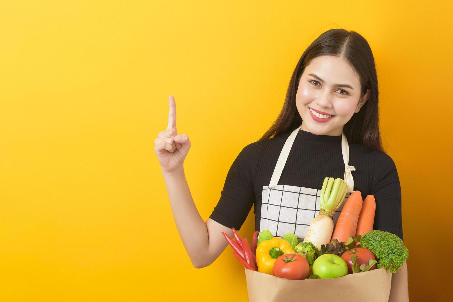 Beautiful young woman is holding vegetables in grocery bag in studio yellow  background photo