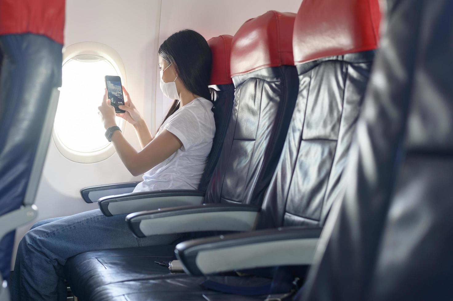 mujer viajera con máscara protectora a bordo del avión usando un teléfono inteligente, viaje bajo la pandemia de covid-19, viajes de seguridad, protocolo de distanciamiento social, nuevo concepto de viaje normal foto