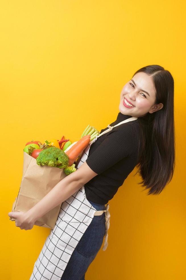 Beautiful young woman is holding vegetables in grocery bag in studio yellow  background photo