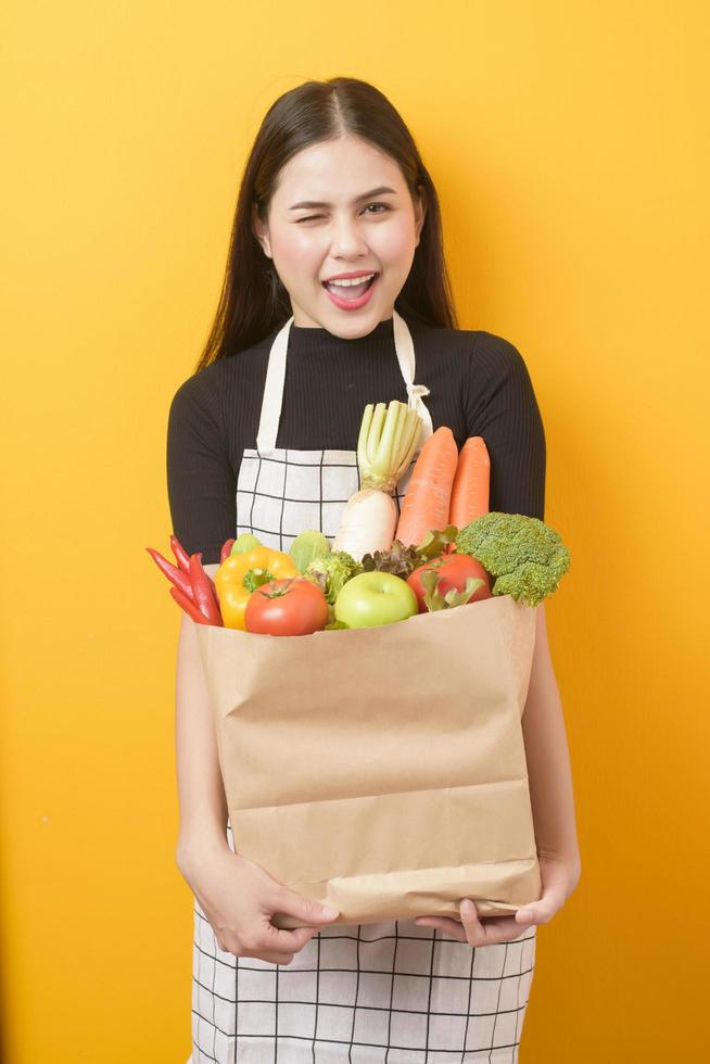 bella joven sostiene verduras en una bolsa de supermercado con fondo amarillo en el estudio foto