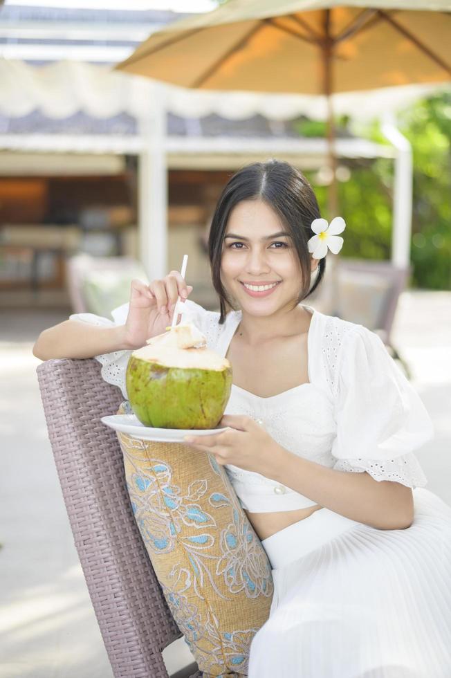 bella mujer turista con flor blanca en el pelo bebiendo coco sentada en un sillón durante las vacaciones de verano foto