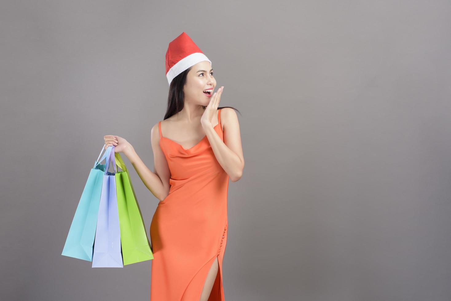 Portrait of young smiling woman wearing red Santa Claus hat is holding credit card and colorful shopping bag isolated gray background studio, Christmas and New Year Concept. photo