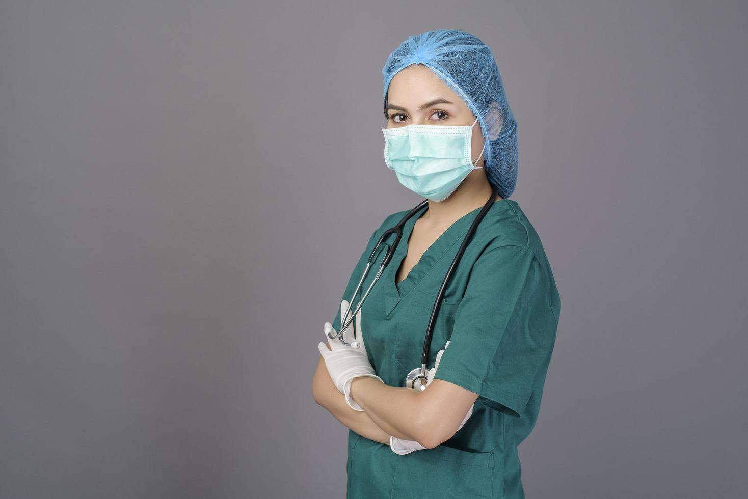 young confident woman doctor in green scrubs is wearing surgical mask over grey background studio photo