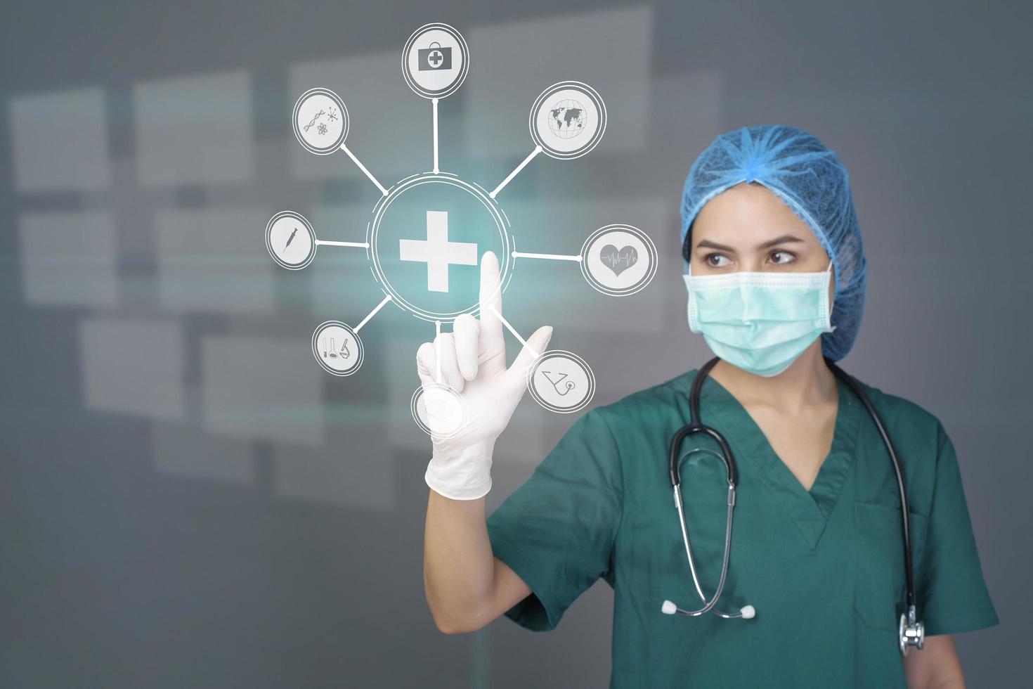 young confident woman doctor in green scrubs is wearing surgical mask over grey background studio , touching medical data on virtual screen , medical technology concept photo