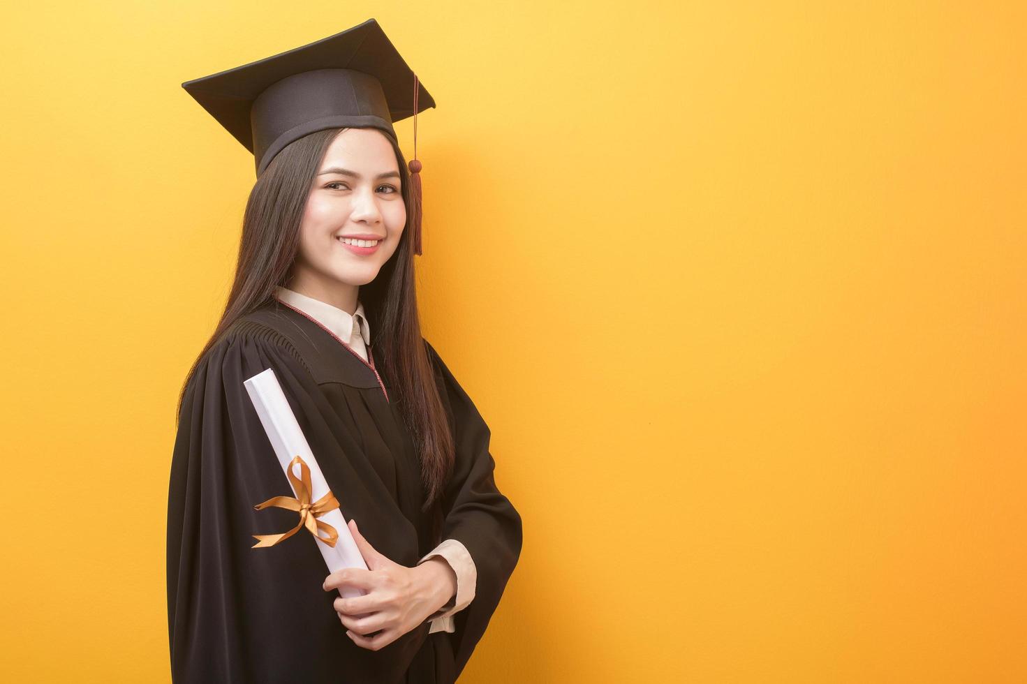 Portrait of happy Beautiful woman in graduation gown is holding education certificate on yellow background photo