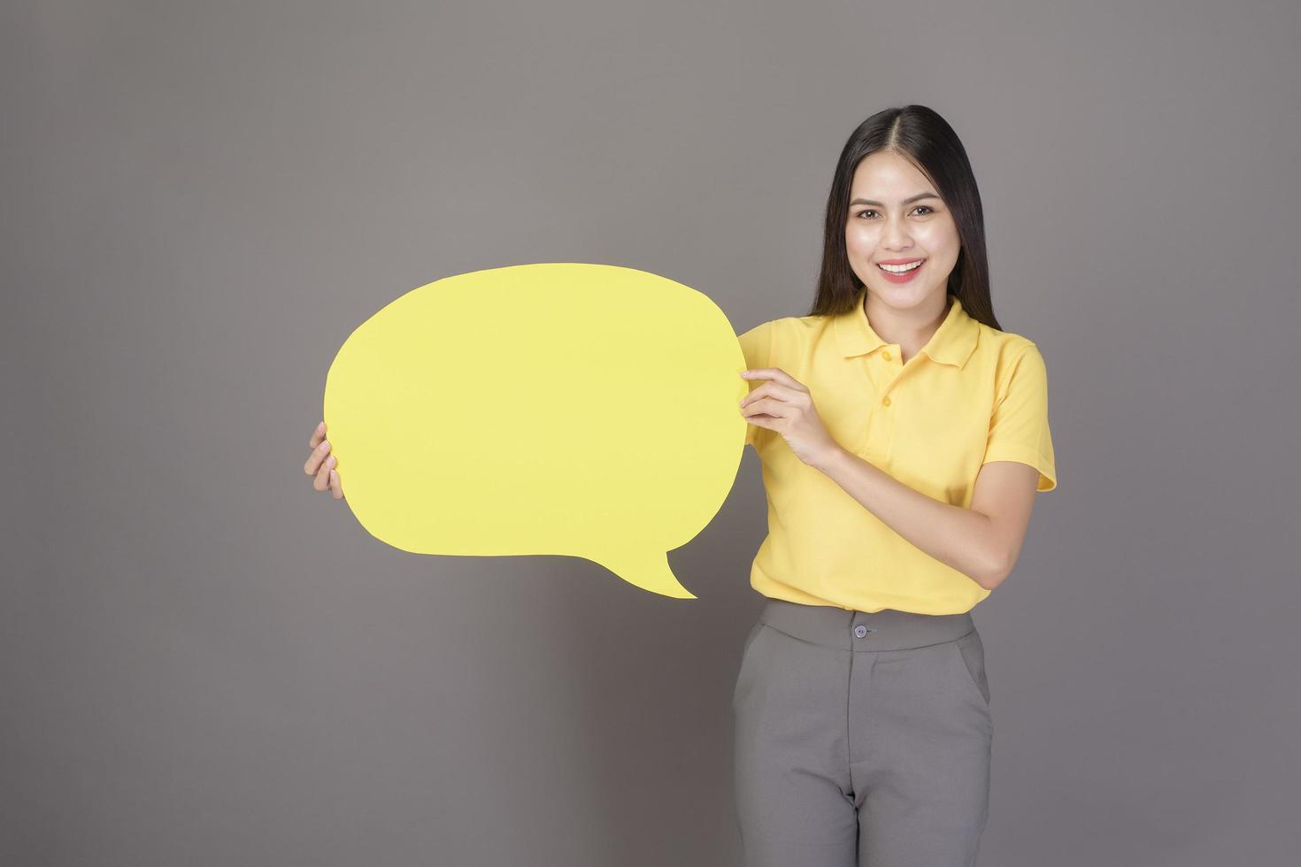 young confident beautiful woman wearing yellow shirt is holding a yellow empty speech on grey background studio photo