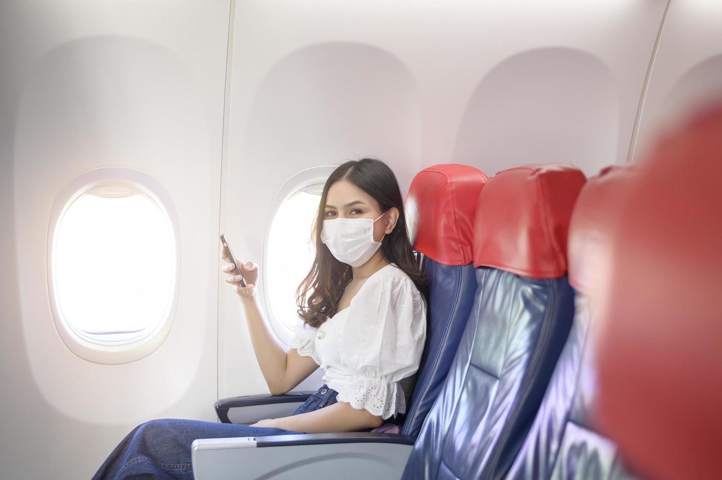 una mujer joven con mascarilla está usando un teléfono inteligente a bordo, nuevo viaje normal después del concepto de pandemia covid-19 foto