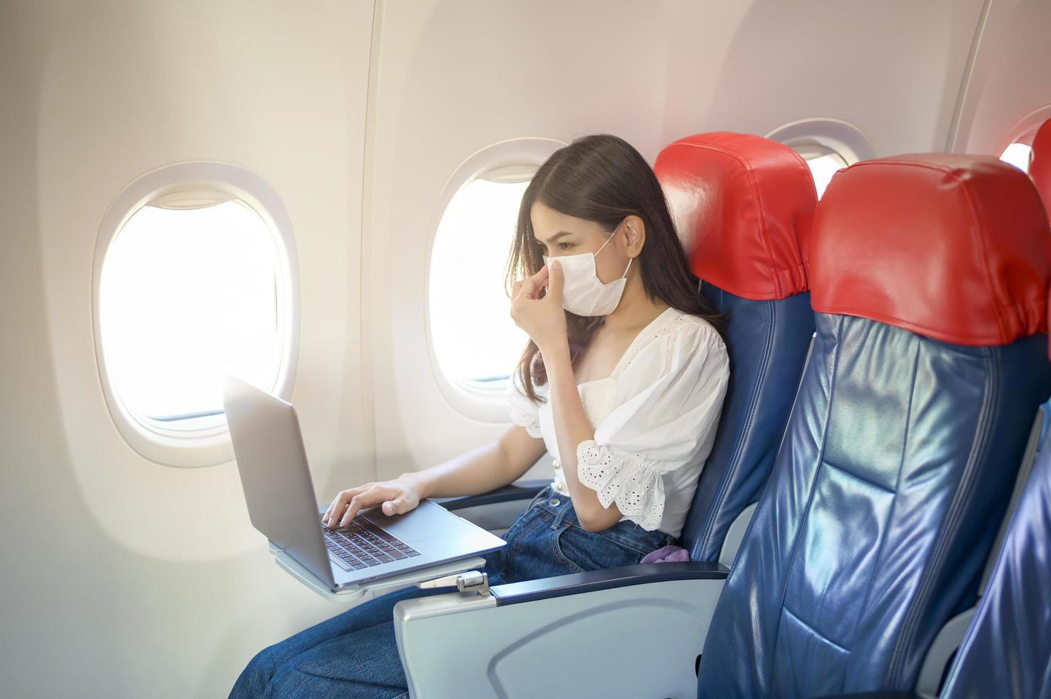 A young woman wearing face mask is using laptop onboard, New normal travel after covid-19 pandemic concept photo