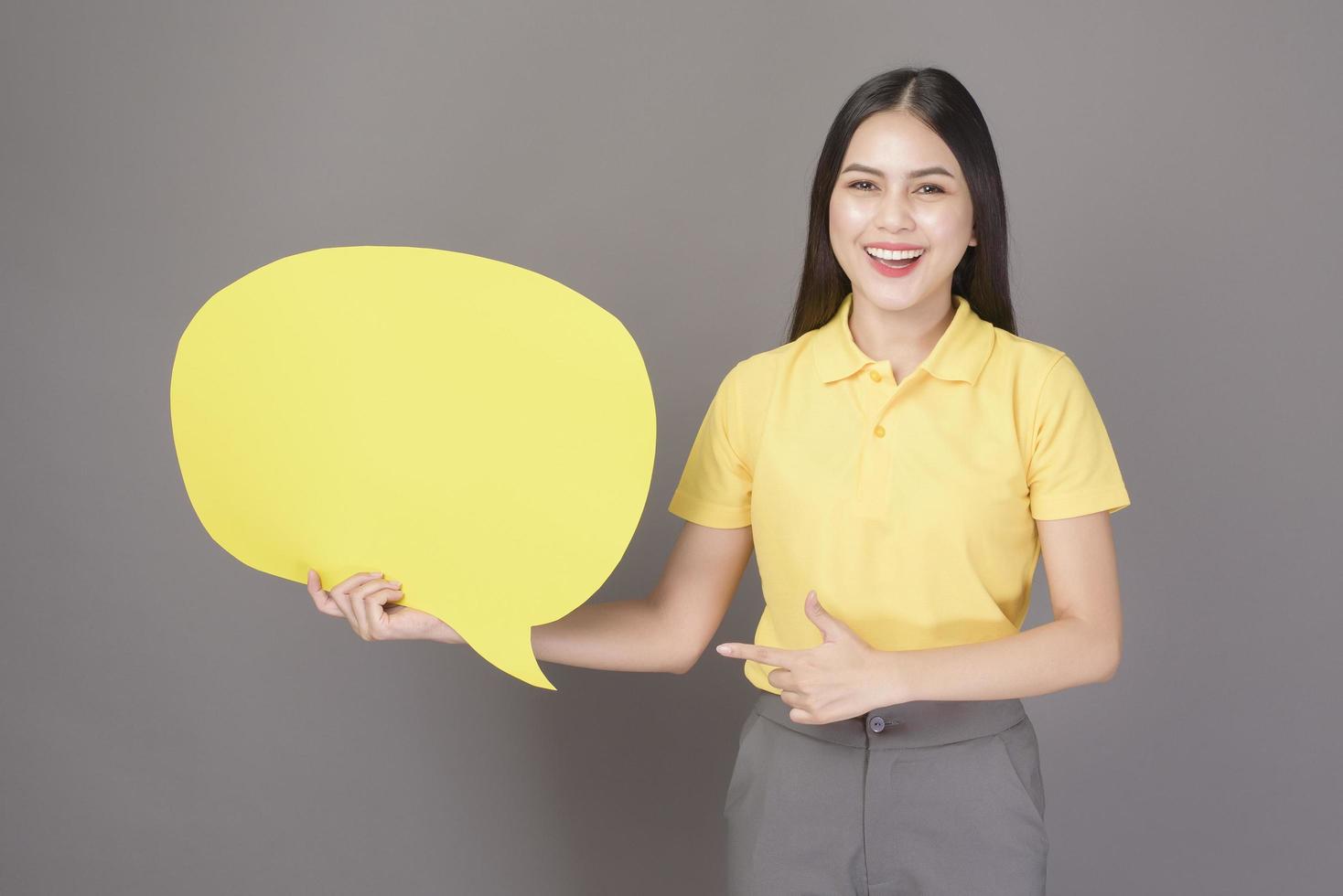 young confident beautiful woman wearing yellow shirt is holding a yellow empty speech on grey background studio photo