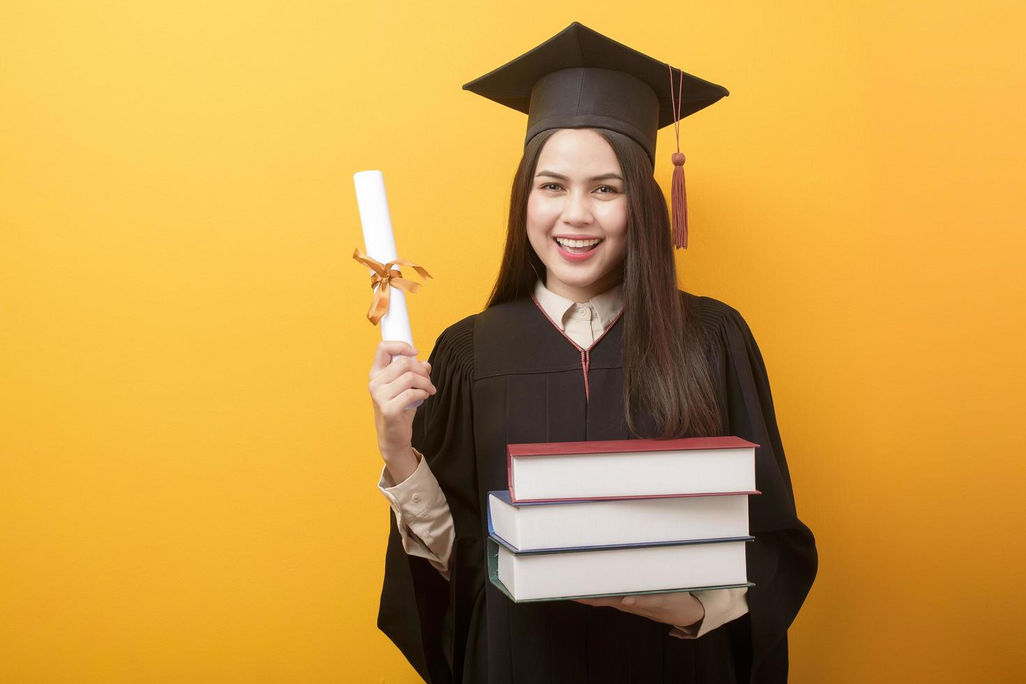 Beautiful woman in graduation gown is holding books and certificate on yellow background photo