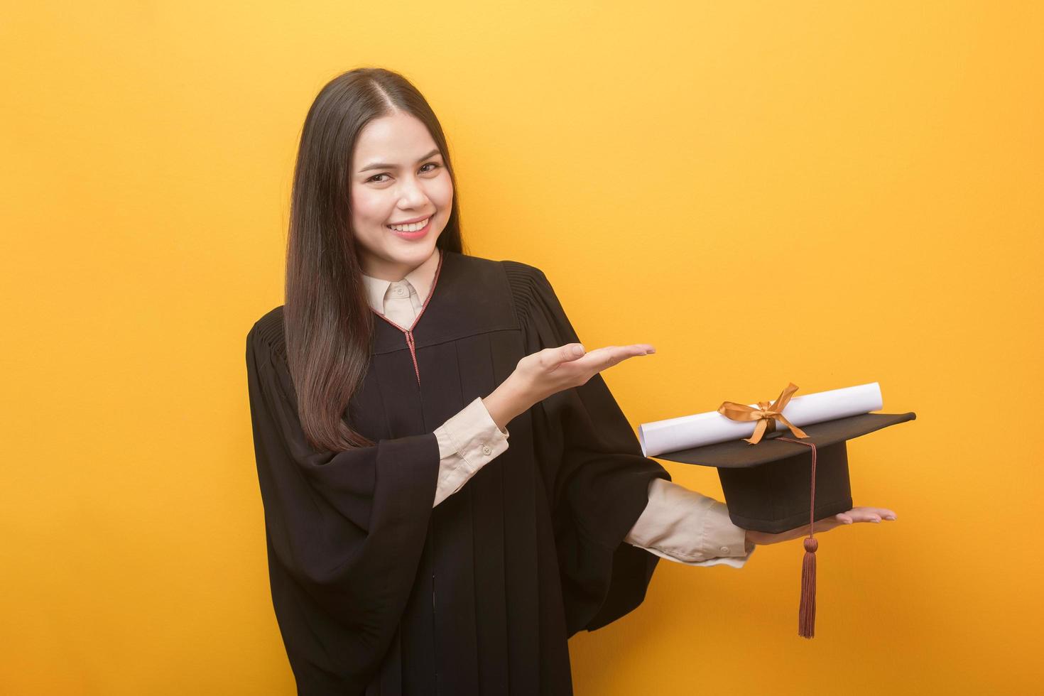 Portrait of happy Beautiful woman in graduation gown is holding education certificate on yellow background photo