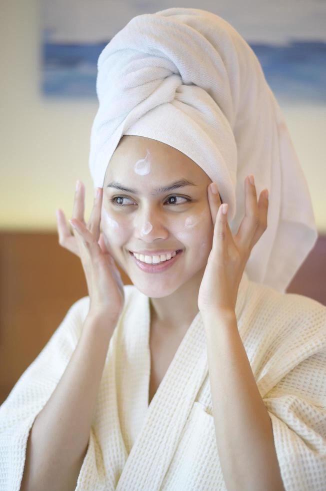 A happy beautiful woman in white bathrobe applying moisturizing cream on face in bedroom, skin care and treatment concept photo