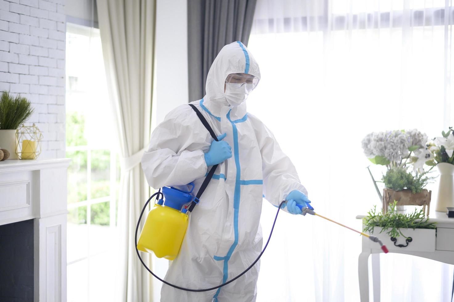 A medical staff in PPE suit is using disinfectant spray in living room, Covid-19 protection , disinfection concept . photo