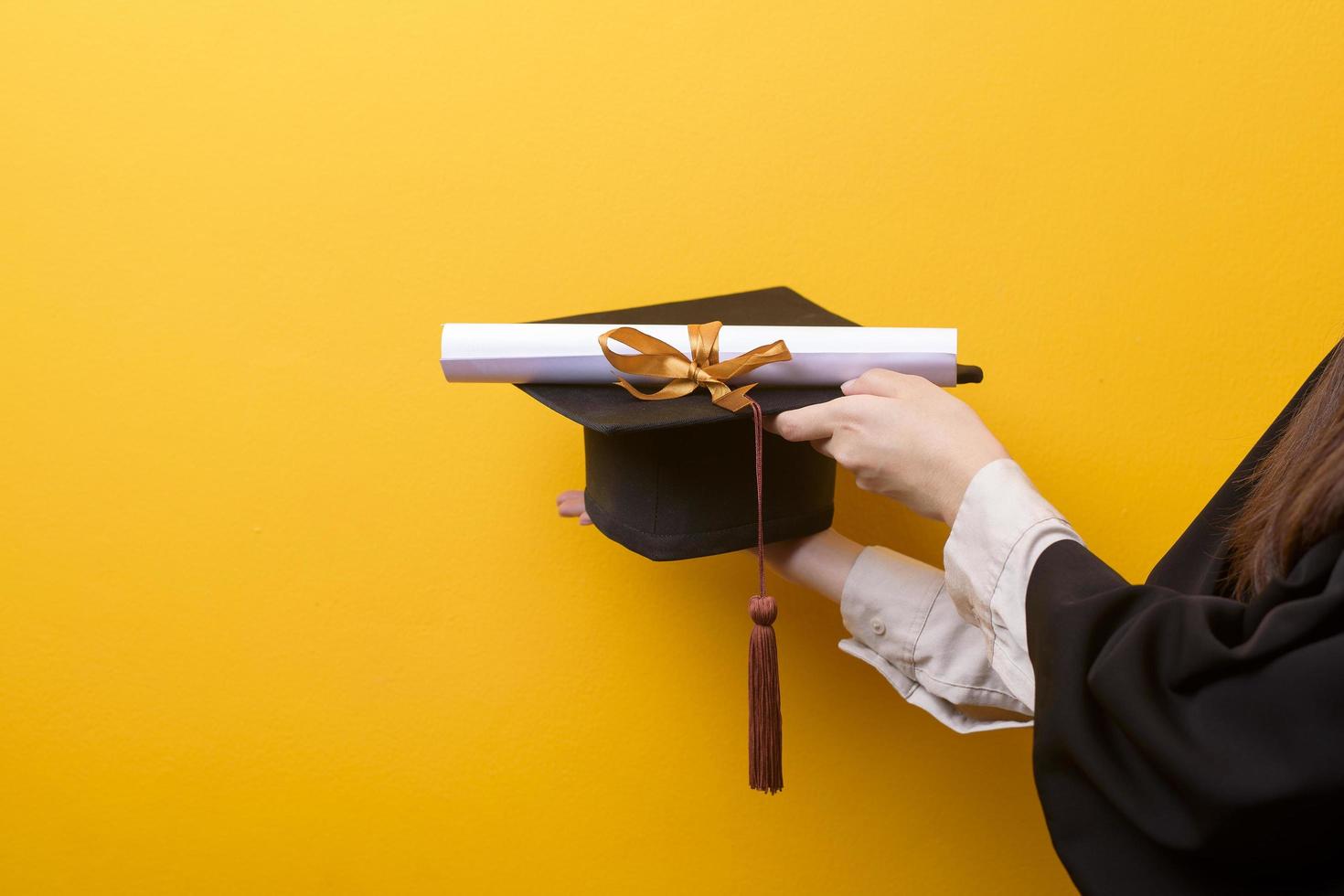 Close up of woman hand in graduation gown is holding graduation cap and certificate on yellow background photo