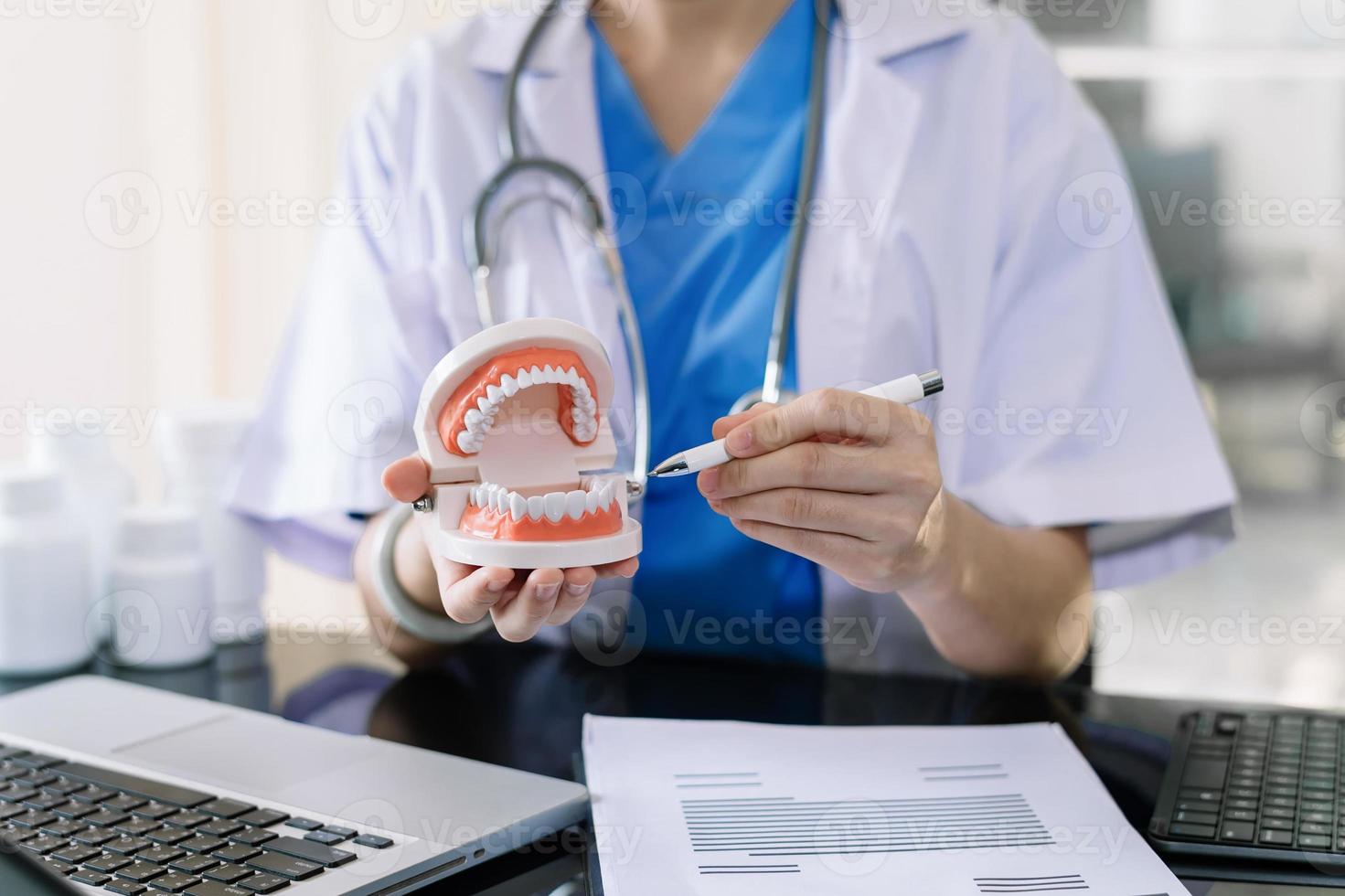 Dentista concentrado sentado a la mesa con muestras de mandíbula modelo de diente y trabajando con tableta y portátil en el consultorio dental foto