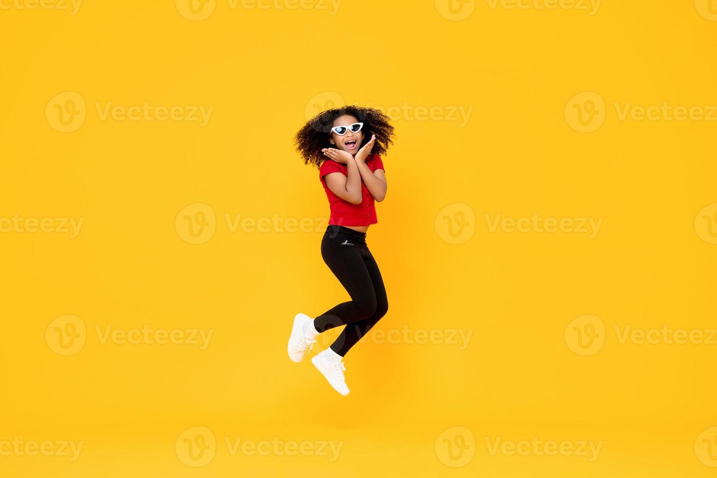 Happy fashionable African American girl jumping in mid-air with hands on chin isolated on yellow background photo