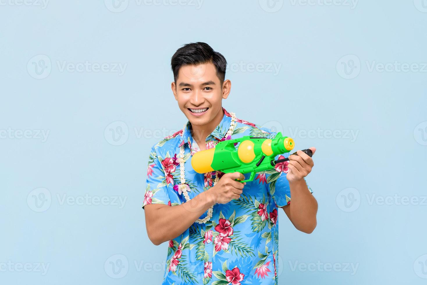 Smiling handsome Asian man playing with water gun isolated on studio blue background for Songkran festival in Thailand and southeast Asia photo