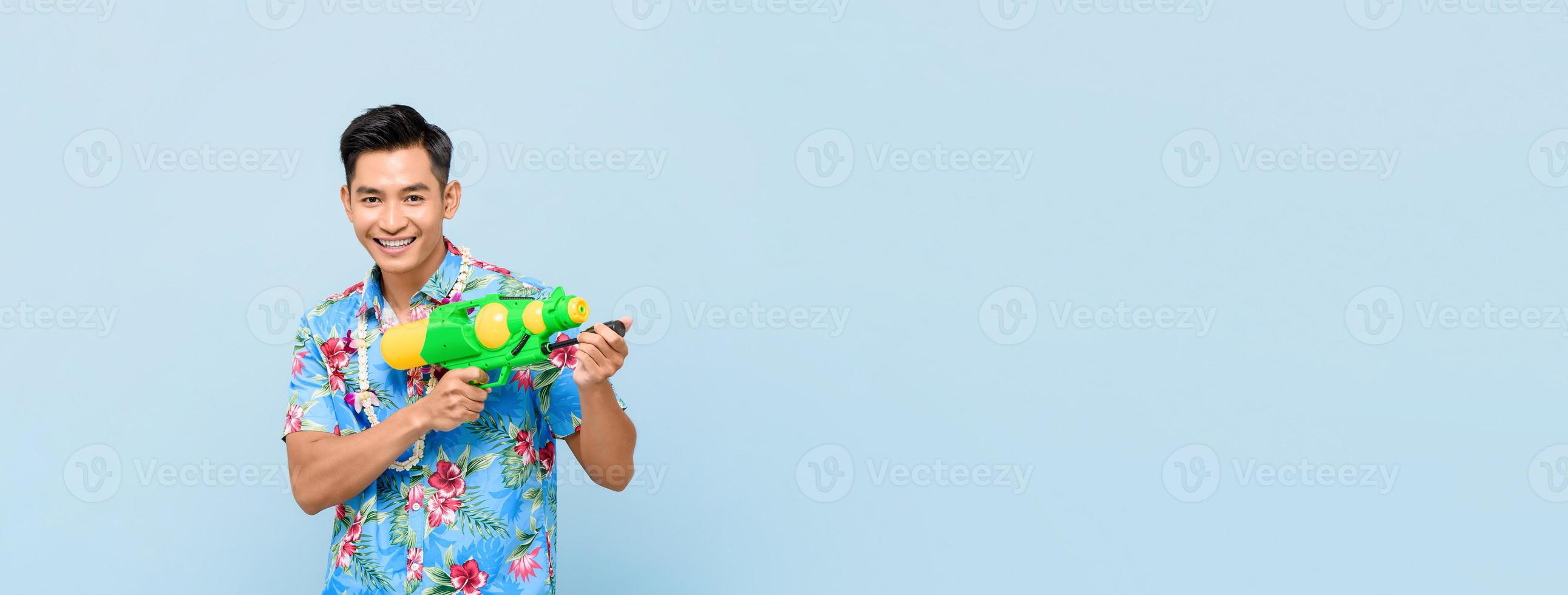 Smiling handsome Asian man playing with water gun isolated on banner blue background with copy space for Songkran festival in Thailand and southeast Asia photo