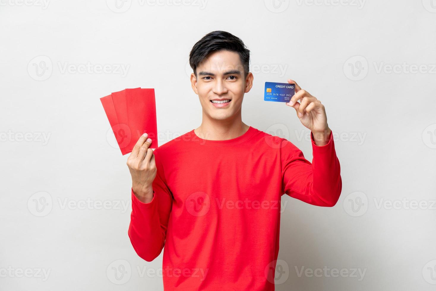 sonriente apuesto hombre asiático sosteniendo sobres rojos o ang pao y tarjeta de crédito en estudio aislado fondo gris claro para conceptos de año nuevo chino foto