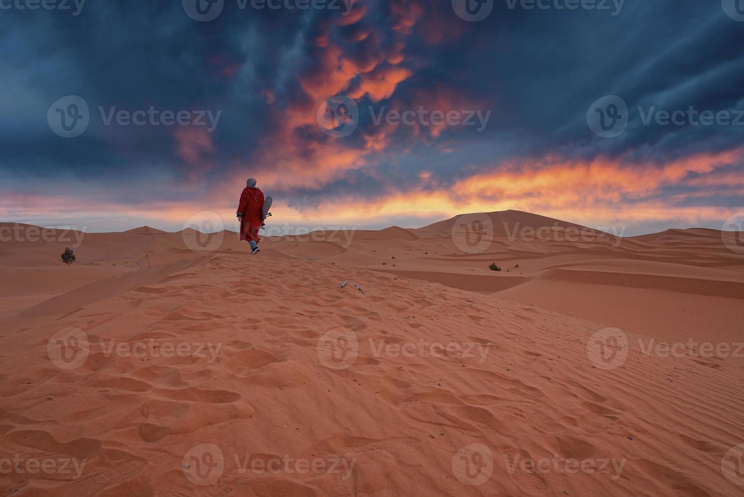 hombre vestido con ropa tradicional con sandboard caminando sobre dunas de arena durante el atardecer foto
