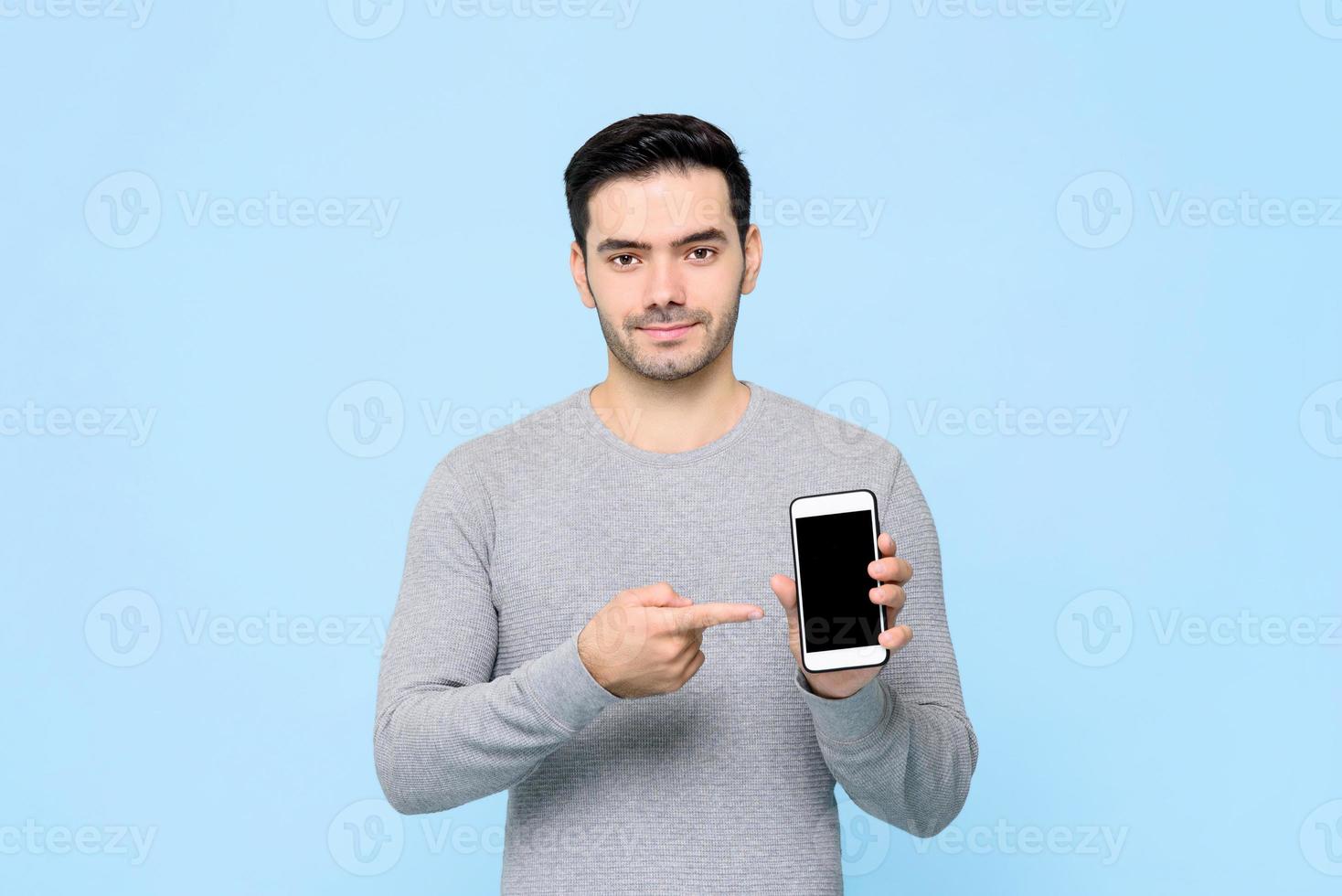 joven apuesto apuntando a su teléfono móvil en la mano aislado en un fondo azul claro foto